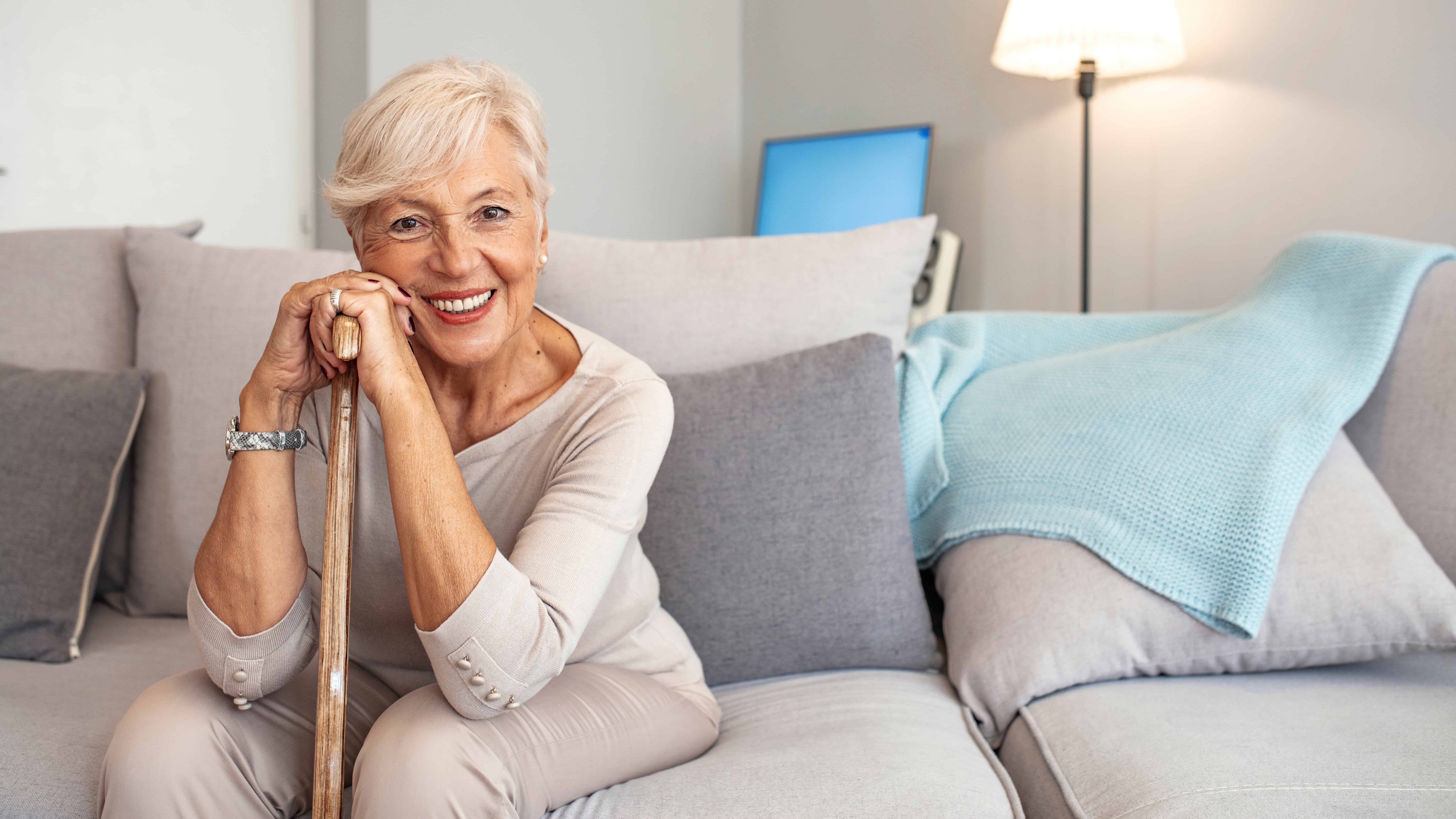 Senior woman smiling with dentures sitting on couch holding cane.
